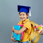 Jeune garçon souriant portant un bonnet de diplômé et un sac à dos rempli de fournitures scolaires pour la rentrée.