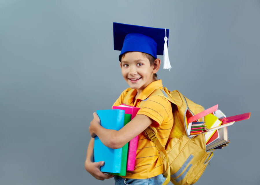 Jeune garçon souriant portant un bonnet de diplômé et un sac à dos rempli de fournitures scolaires pour la rentrée.