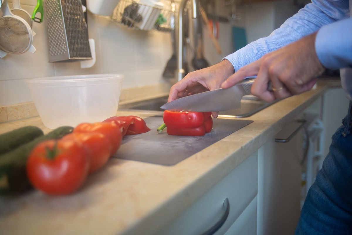 Personnage tranchant un poivron rouge sur une planche à découper, tomates et concombres à proximité