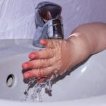 Petite main d'enfant en rose se lavant sous un robinet dans un lavabo blanc