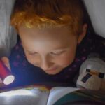 Enfant sous couette, lampe torche éclaire un livre ouvert
